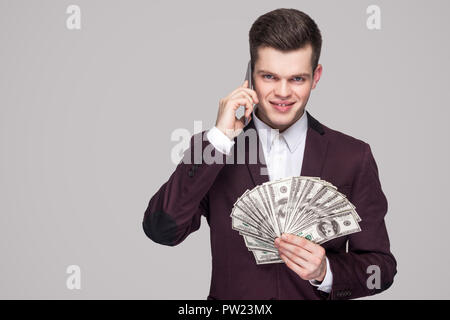 Portrait of attractive young businessman prospère en manteau violet classique debout, montrant fan de l'argent et faire des arrangements sur téléphone mobile. Indoo Banque D'Images