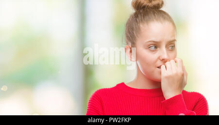 Jeune femme blonde portant chignon et pull rouge à souligné et nerveux avec les mains sur la bouche de mordre les ongles. Problème d'anxiété. Banque D'Images