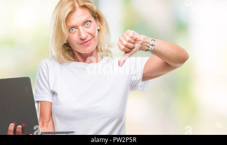 L'âge moyen à l'aide d'un ordinateur portable femme blonde au visage en colère avec fond isolé, signe négatif montrant non avec Thumbs down, rejet conc Banque D'Images
