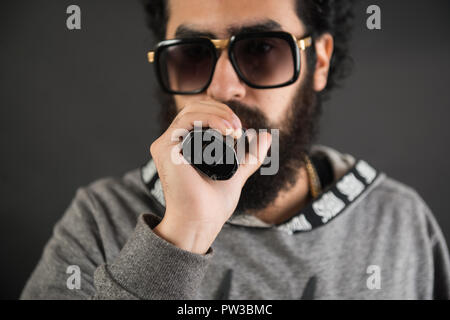 Portrait d'un jeune mec avec grande barbe et des lunettes de soleil et un vaping cigarette électronique sur un fond noir. Banque D'Images