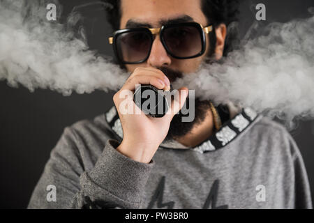 Portrait d'un jeune mec avec grande barbe et des lunettes de soleil et un vaping cigarette électronique sur un fond noir. Banque D'Images