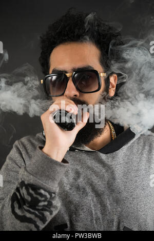 Portrait d'un jeune mec avec grande barbe et des lunettes de soleil et un vaping cigarette électronique sur un fond noir. Banque D'Images