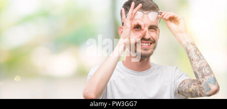 Les jeunes adultes faisant l'homme tatoué ok geste avec la main en souriant, les yeux regardant à travers les doigts avec un visage heureux. Banque D'Images