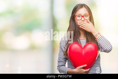 Young Asian Woman in love holding lire plus coeur fond isolé couvrir la bouche à part choqué avec honte pour erreur, expression de la peur, peur Banque D'Images