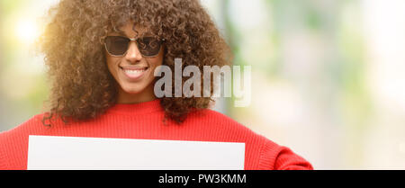 African American Woman wearing sunglasses holding blank bandeau publicitaire, bonne affiche pour votre annonce, offre ou annonce, grand panneau de papier Banque D'Images