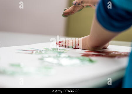Closeup portrait of toddler enfant faisant colourfull palmaires avec les peintures au doigt sur un morceau de papier blanc. Banque D'Images