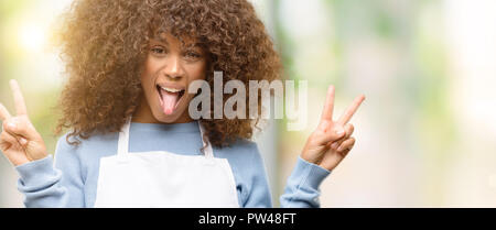 African American boutique femme portant un tablier, looking at camera montrant la langue et faire de la victoire avec les doigts Banque D'Images