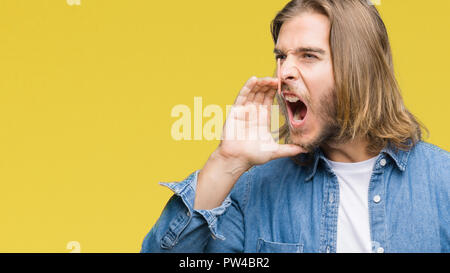Jeune homme séduisant aux cheveux longs sur fond isolé crier et crier fort à côté avec la main sur la bouche. Concept de communication. Banque D'Images