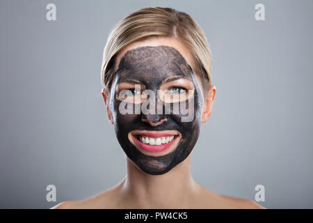 Souriante Jeune femme avec du charbon activé Masque de visage à l'arrière-plan gris Banque D'Images