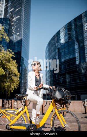 Portrait d'une femme d'affaires élégant en costume blanc debout avec vélo à le quartier financier avec les bâtiments modernes sur l'arrière-plan Banque D'Images