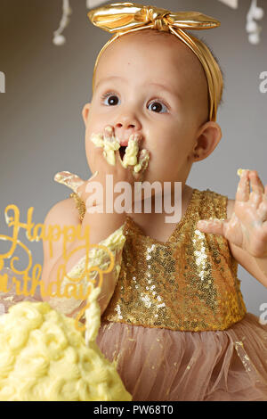 Petite fille Bébé doux dans une robe d'or avec un arc sur la tête d'essayer un gâteau de gelée jazzy une crème. studio shot of un anniversaire sur fond gris entouré par des boules Banque D'Images