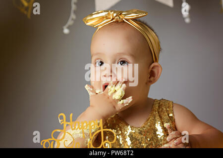 Petite fille Bébé doux dans une robe d'or avec un arc sur la tête d'essayer un gâteau de gelée jazzy une crème. studio shot of un anniversaire sur fond gris entouré par des boules Banque D'Images