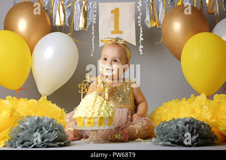 Une petite fille douce, dans une robe d'or avec un arc sur la tête d'essayer un gâteau de gelée jazzy une crème. studio shot of un anniversaire sur fond gris entouré par des boules Banque D'Images