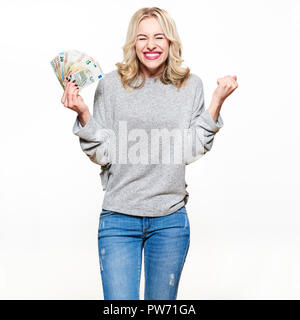 Super excitée jeune femme en jeans et pull gris holding bunch des billets en euros, le clinchage poings, gain de loterie célébration. Ecstatic woman hold Banque D'Images