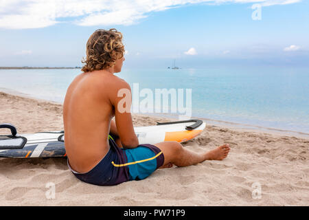 Bel homme est assis sur la plage avec un voile blanc vide attendre Vague à surf en mer ocean shore. Concept de sport, fitness, liberté, bonheur, nouvelle vie moderne, hipster. Banque D'Images