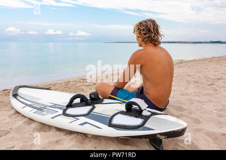 Bel homme est assis sur la plage avec un voile blanc vide attendre Vague à surf en mer ocean shore. Concept de sport, fitness, liberté, bonheur, nouvelle vie moderne, hipster. Banque D'Images