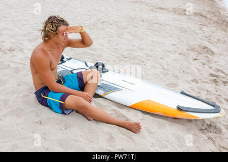 Bel homme est assis sur la plage avec un voile blanc vide attendre Vague à surf en mer ocean shore. Concept de sport, fitness, liberté, bonheur, nouvelle vie moderne, hipster. Banque D'Images