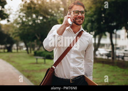 Homme portant un sac de bureau de conversations au téléphone portable en marche de bureau. Smiling businessman allant à l'office de tourisme à pied tôt le matin. Banque D'Images
