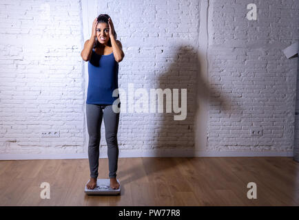 Jeune femme slim fit et contrôle de poids du corps sur l'échelle se trouvant en gras et se sentir déprimé et désespéré avec grande lumière ombre edgy en mangeant di Banque D'Images