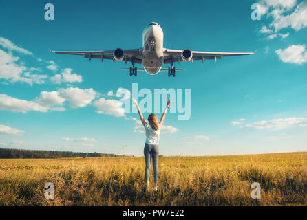 Avion et femme au coucher du soleil. Paysage d'automne avec fille debout sur la pelouse avec les armes et le vol de l'avion de passagers en bleu ciel. Wo Banque D'Images
