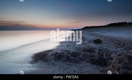 Coucher de soleil sur la mer Baltique avec rivage allant dans l'horizon. Le sable et les vagues lisses et soyeux avec des forêts d'arbres à l'horizon. Banque D'Images