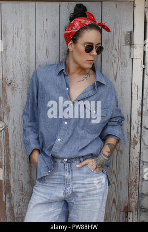 La mode femme Cool hipster avec des lunettes avec un tatouage sur les mains d'un denim shirt et jeans vintage avec un fichu rouge se tient près d'un mur en bois Banque D'Images