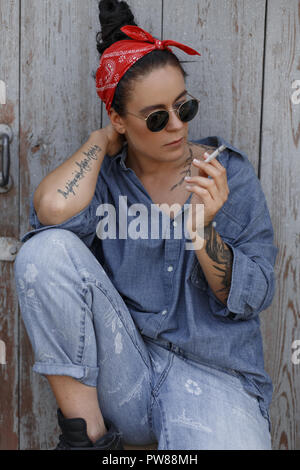 Belle américaine élégante femme denim hipster dans des vêtements à la mode avec un fichu rouge et lunettes de fumer une cigarette et assis près de un sol en bois Banque D'Images