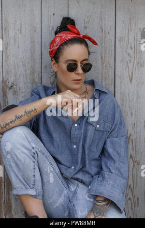 American Cool hipster femme élégante avec des lunettes d'un denim shirt et jeans fashion avec une cigarette est assis près d'un vieux mur en bois. Style Pin Up Banque D'Images