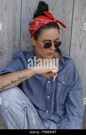 Cool belle américaine à la mode avec des lunettes de soleil femme modèle hipster en bleu vintage denim jeans élégant vêtements et assis près d'un mur en bois Banque D'Images