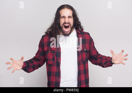 Bel homme en colère avec barbe et black long cheveux bouclés dans un style décontracté, chemise rouge à carreaux standing looking at camera et crier avec les bras levés. Banque D'Images