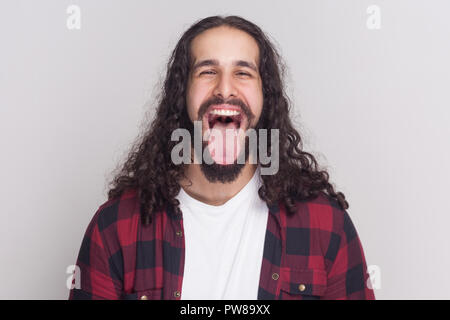 Crazy bel homme avec barbe et cheveux longs noirs dans un style décontracté, chemise rouge à carreaux, permanent ouvrir grand la bouche, de la langue et à la recherche au camer Banque D'Images