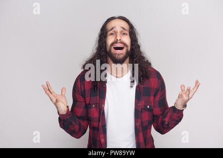 Ce que je dois faire ? Triste Malheureux homme avec barbe et black long cheveux bouclés en chemise rouge à carreaux debout avec les bras levés, à la caméra et à poser Banque D'Images