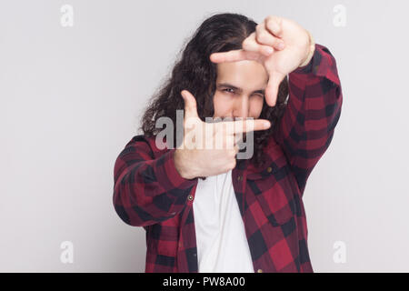 Portrait de bel homme avec barbe et cheveux longs noirs dans un style décontracté, chemise rouge à carreaux et à rendre permanent le châssis de l'appareil photo avec les mains Banque D'Images