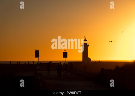 Un groupe de personnes se tiennent près de un phare avant le coucher du soleil avec vol de mouettes par sur le ciel d'or. Banque D'Images