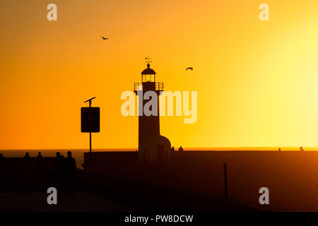 Un groupe de personnes se tiennent près de un phare avant le coucher du soleil avec vol de mouettes par sur le ciel d'or. Banque D'Images