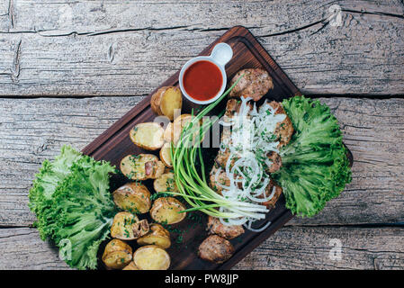 Morceaux de porc grillé appétissant sur le grill, présenté sur une planche en bois, ainsi que des feuilles de salade verte et de pommes de terre avec la sauce tomate. Banque D'Images