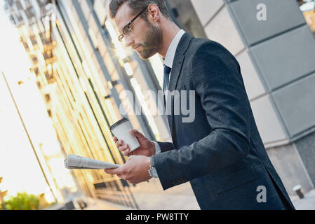 Jeune homme en costume et lunettes tenant une tasse de papier et la lecture de journaux d'affaires dans ses mains Banque D'Images