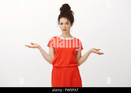 Portrait d'indifférent careless portrait femme aux cheveux bouclés en robe rouge, épaules avec les mains et se propager à regarder la caméra, remis en cause et pas au courant, question d'être dérangé avec Banque D'Images