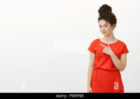 Portrait of creative charmante femme aux cheveux bouclés en étudiant avec robe rouge tendance coiffure chignon, pointant dans le coin en haut à gauche et levage sourcil tout en suggérant quelque chose ou qui ont une grande idée Banque D'Images