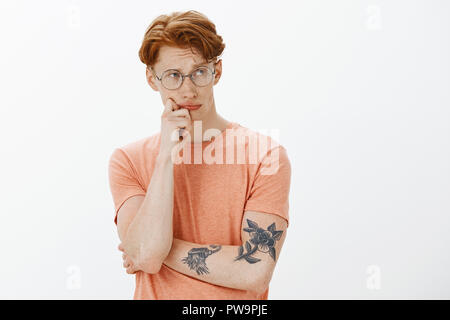 Moody et réfléchie des hommes jeunes européens aux cheveux roux et taches de rousseur, fixant droit sérieux et ciblé, toucher et à lèvre froncer, ce choix important ou de décision sur fond gris Banque D'Images