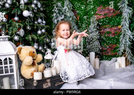 Jolie petite fille en robe blanche assis près d'un arbre de Noël sur une valise à côté de l'une des bougies et des ours en peluche et projette la neige jusqu' Banque D'Images