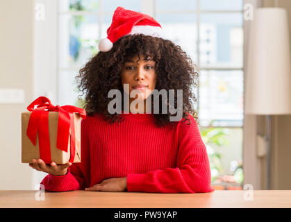 African American Woman holding present wearing christmas red hat avec une expression confiante face à la pensée intelligente grave Banque D'Images