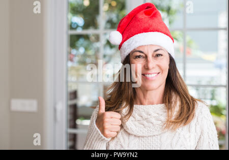 Young woman wearing santa claus hat à la maison heureux avec un grand sourire faisant signe ok, pouce vers le haut avec les doigts, signe d'excellentes Banque D'Images