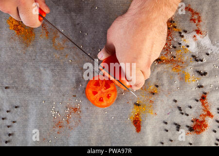 Gros plan du male chef coupe manuelle une tomate rouge mûre avec un couteau de cuisine dans le contexte d'une table de cuisine avec des épices, sel, poivre, vue du dessus Banque D'Images