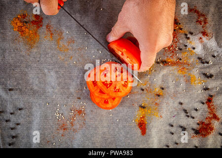 Gros plan du male chef coupe manuelle une tomate rouge mûre avec un couteau de cuisine dans le contexte d'une table de cuisine avec des épices, sel, poivre, vue du dessus Banque D'Images