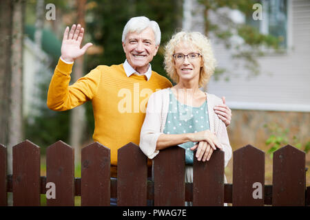 Hospitalier mature couple standing by fence tout en agitant la main de l'homme sympathique à leurs voisins Banque D'Images