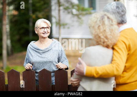 Femme mature blonde à lunettes et pull gris en regardant ses voisins au cours de parler par fence Banque D'Images