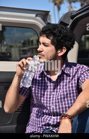 Un beau jeune homme à l'attitude de l'eau en bouteille de boisson sur la porte arrière de sa voiture. Banque D'Images