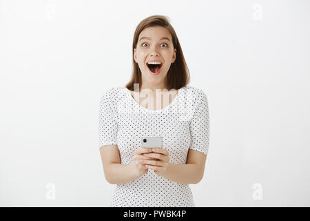 La technologie, les émotions et le style de concept. Portrait de charmant impressionnée et happy cute woman, exprimant sa surprise, d'être heureux alors que tout sourire à l'appareil photo, holding smartphone sur mur gris Banque D'Images