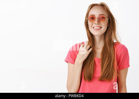 Studio shot of cute heureux et emotive jeune rousse femelle en lunettes roses et t-shirt de toucher mèche de cheveux et se sentir à l'aise en souriant société inconnu Essayer démarrer la conversation friendly Banque D'Images